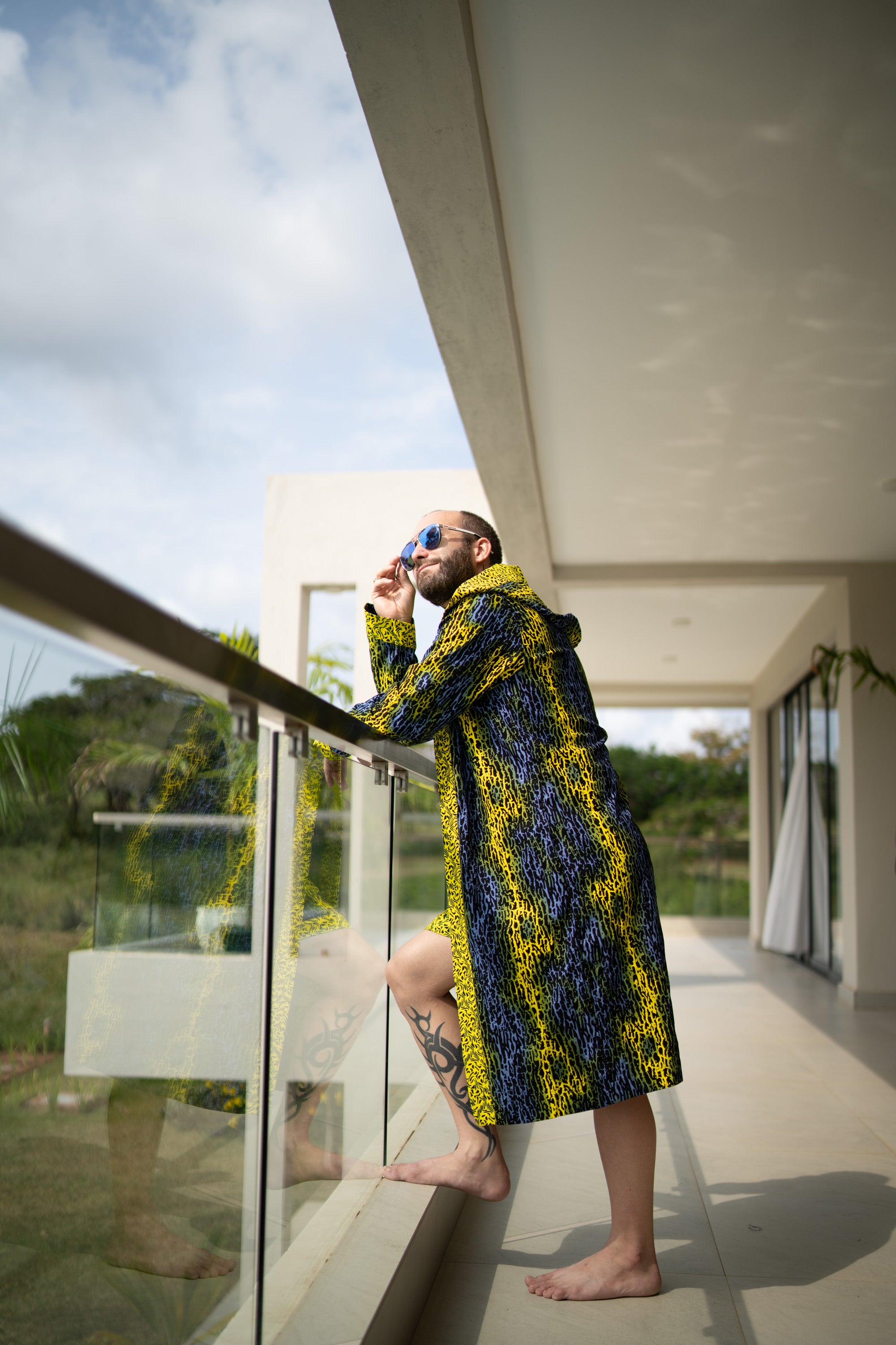 A man styling Electric Safari African cover-up and shorts set in safari-inspired print – Zawadi Hat  on a balcony with a modern building and greenery in the background.
