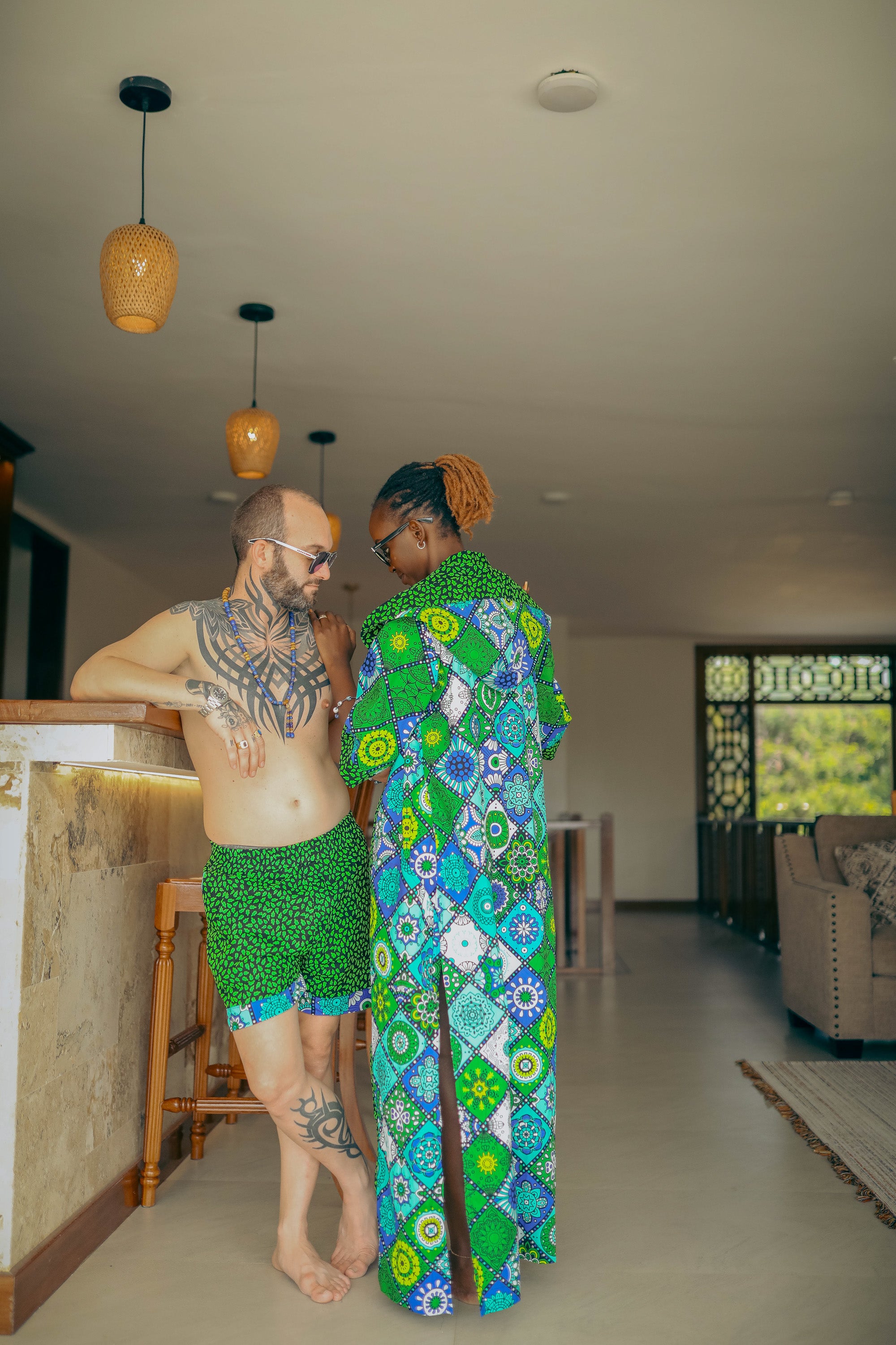 A couple wearing green and patterned matching outfits in a room with pendant lights and a couch.
