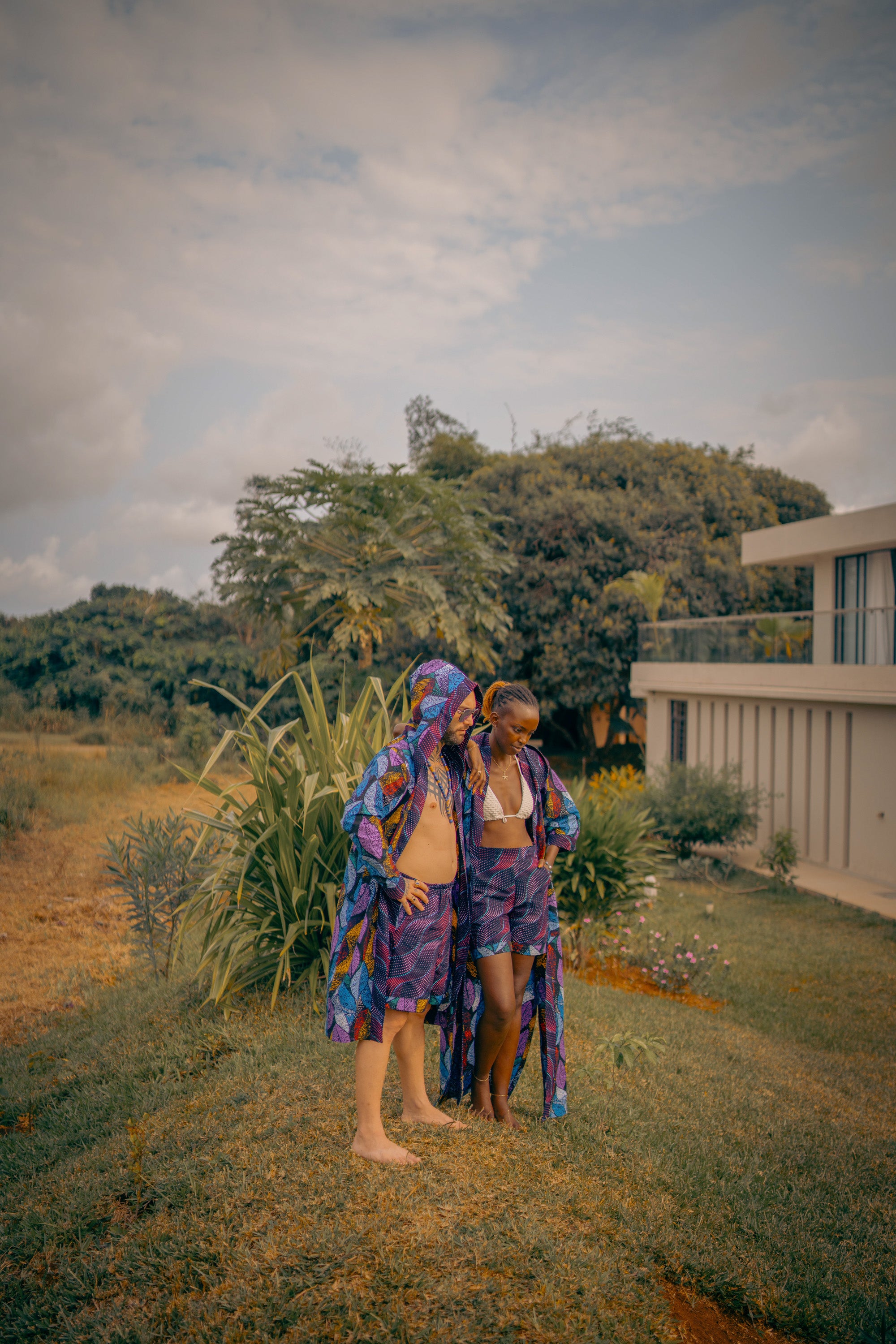 A  couple wrapped in colorful kimonos standing in a garden with trees and a building in the background with sunset light.
