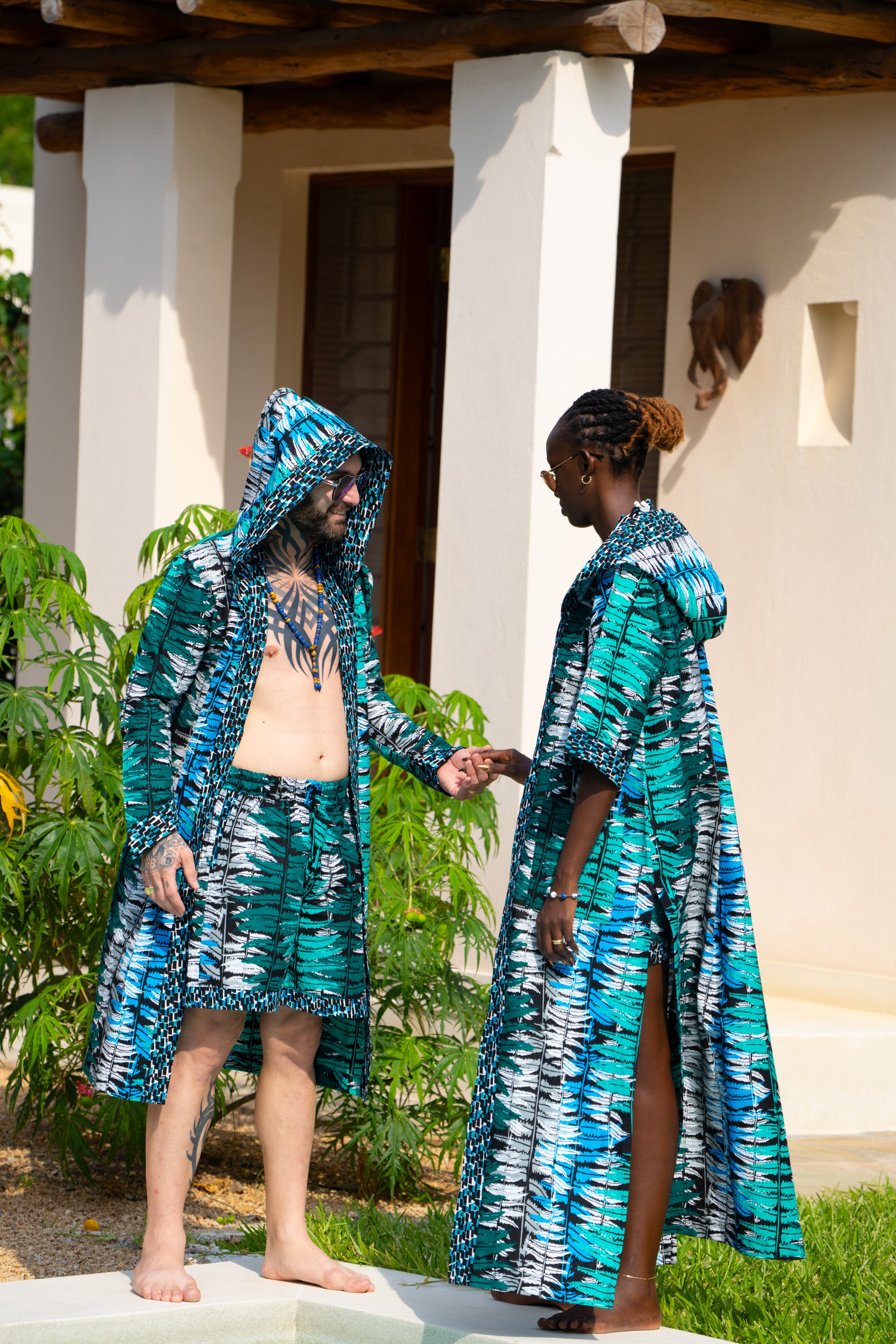 A couple in Ocean Teal African cover-up and shorts set standing by a pool with a building and plants in the background.