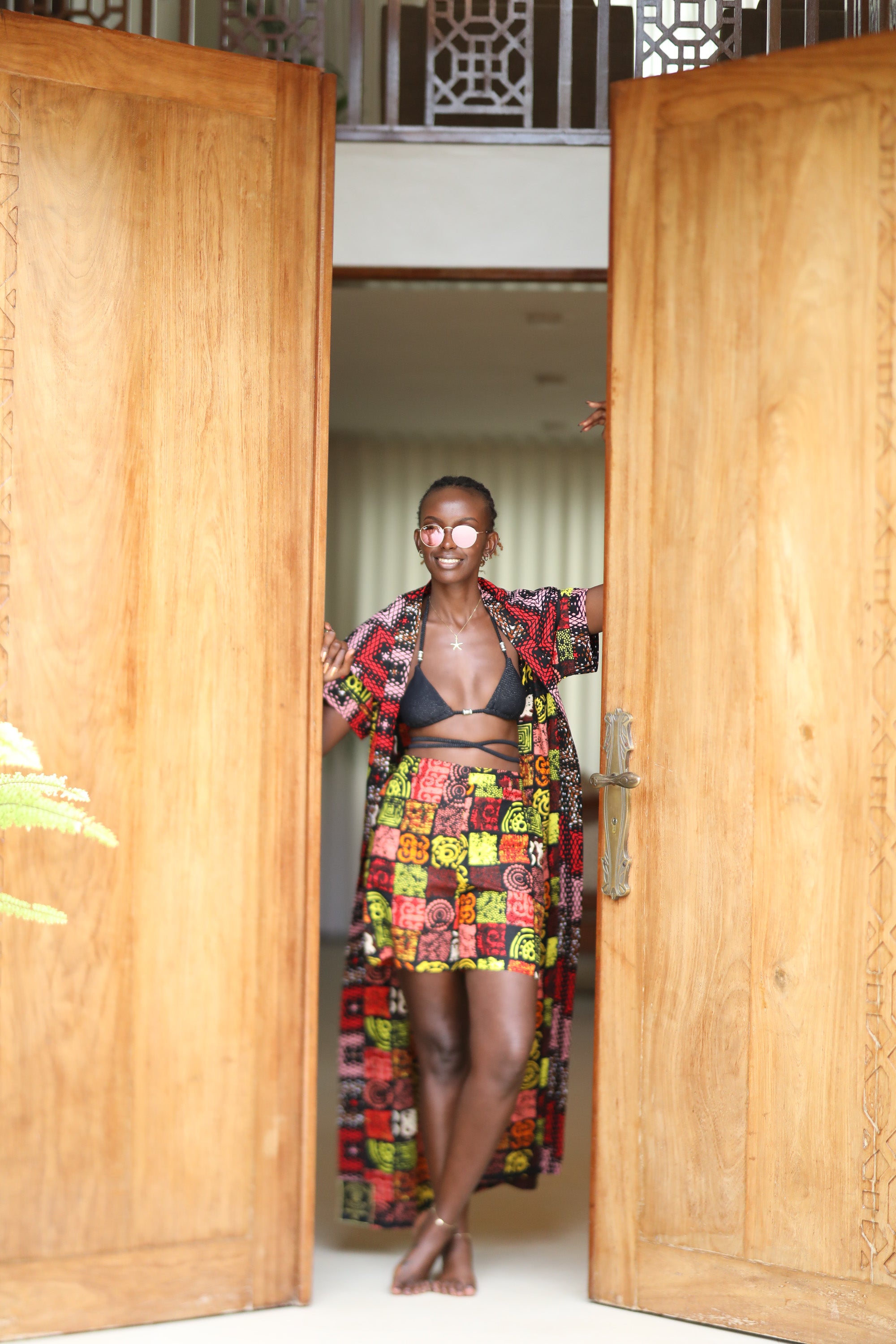Woman styling Electric Safari African cover-up and shorts set in safari-inspired print – Zawadi Hat Fashion standing in an open wooden door.
