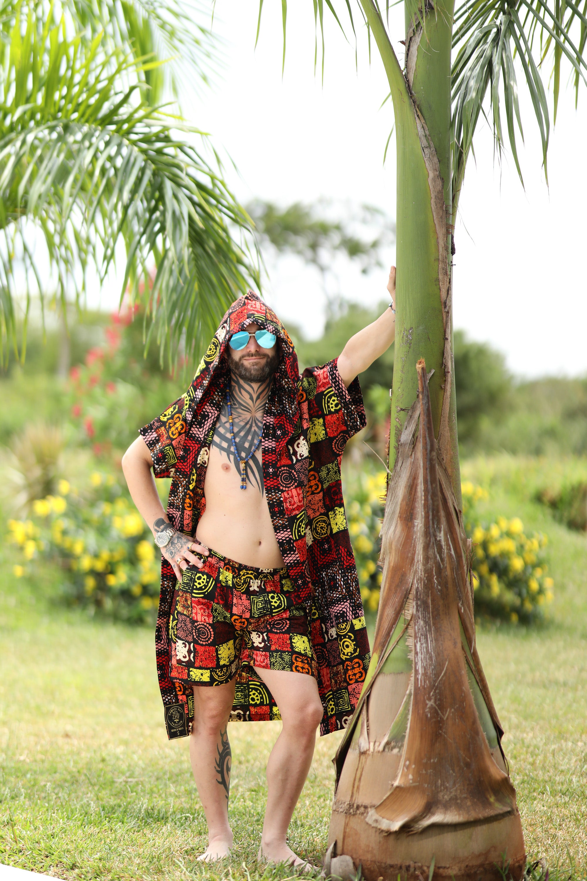 A man styling Electric Safari African cover-up safari-inspired print – Zawadi Hat standing next to a palm tree in a garden setting