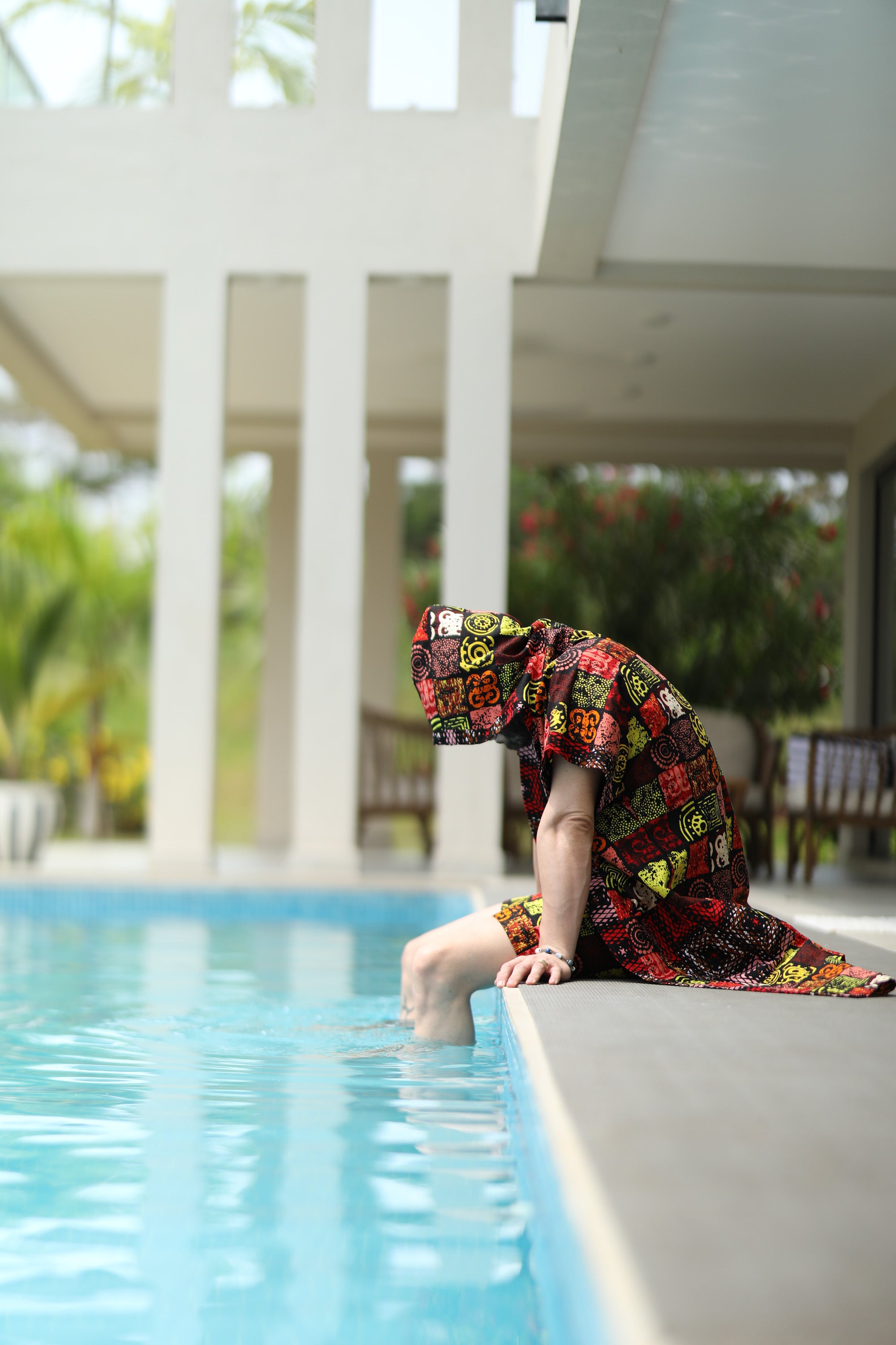 A man styling Electric Safari African cover-up safari-inspired print – Zawadi Hat sitting by a pool with a tropical setting.