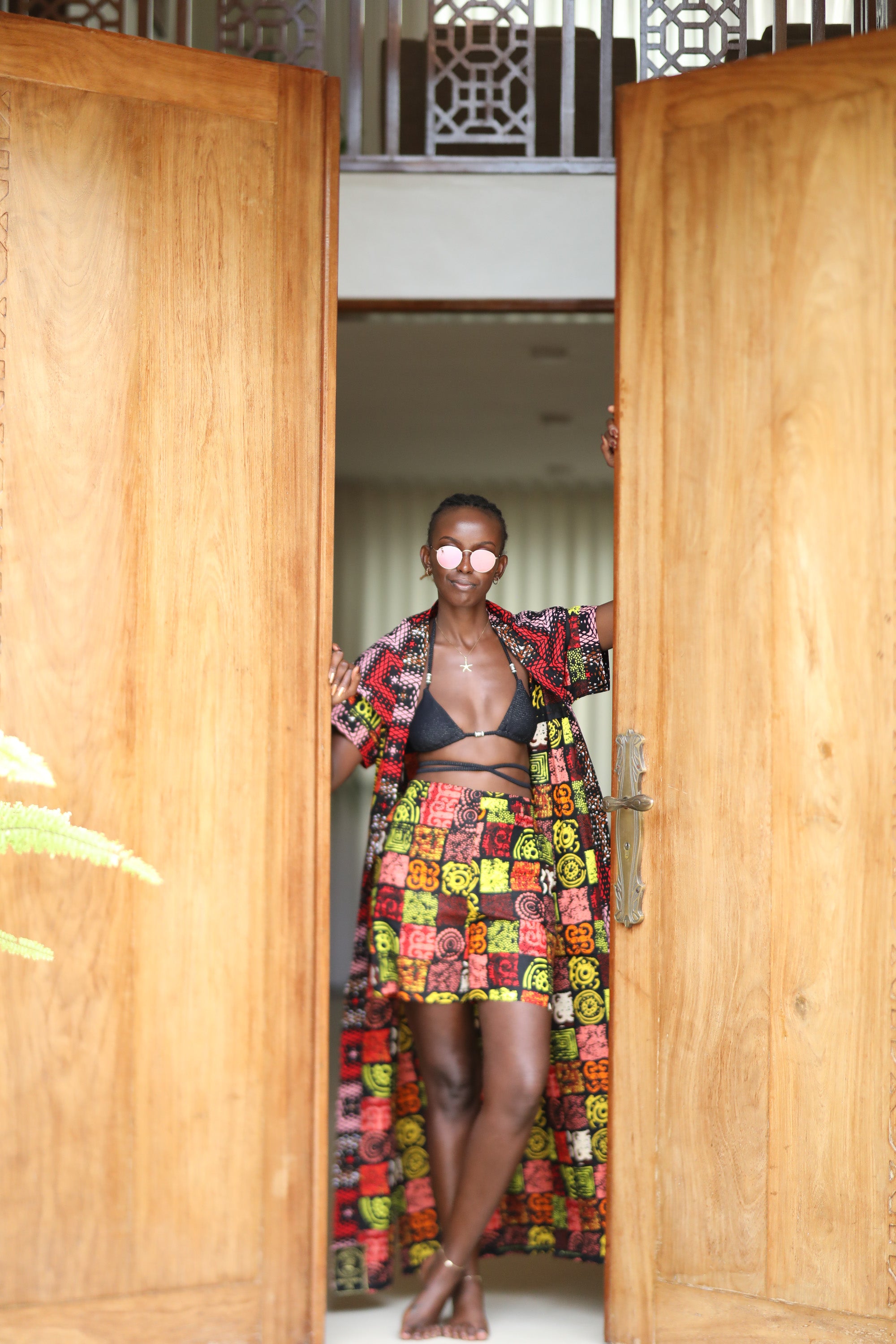 Woman styling Electric Safari African cover-up and shorts set in safari-inspired print – Zawadi Hat Fashion standing in an open wooden door.