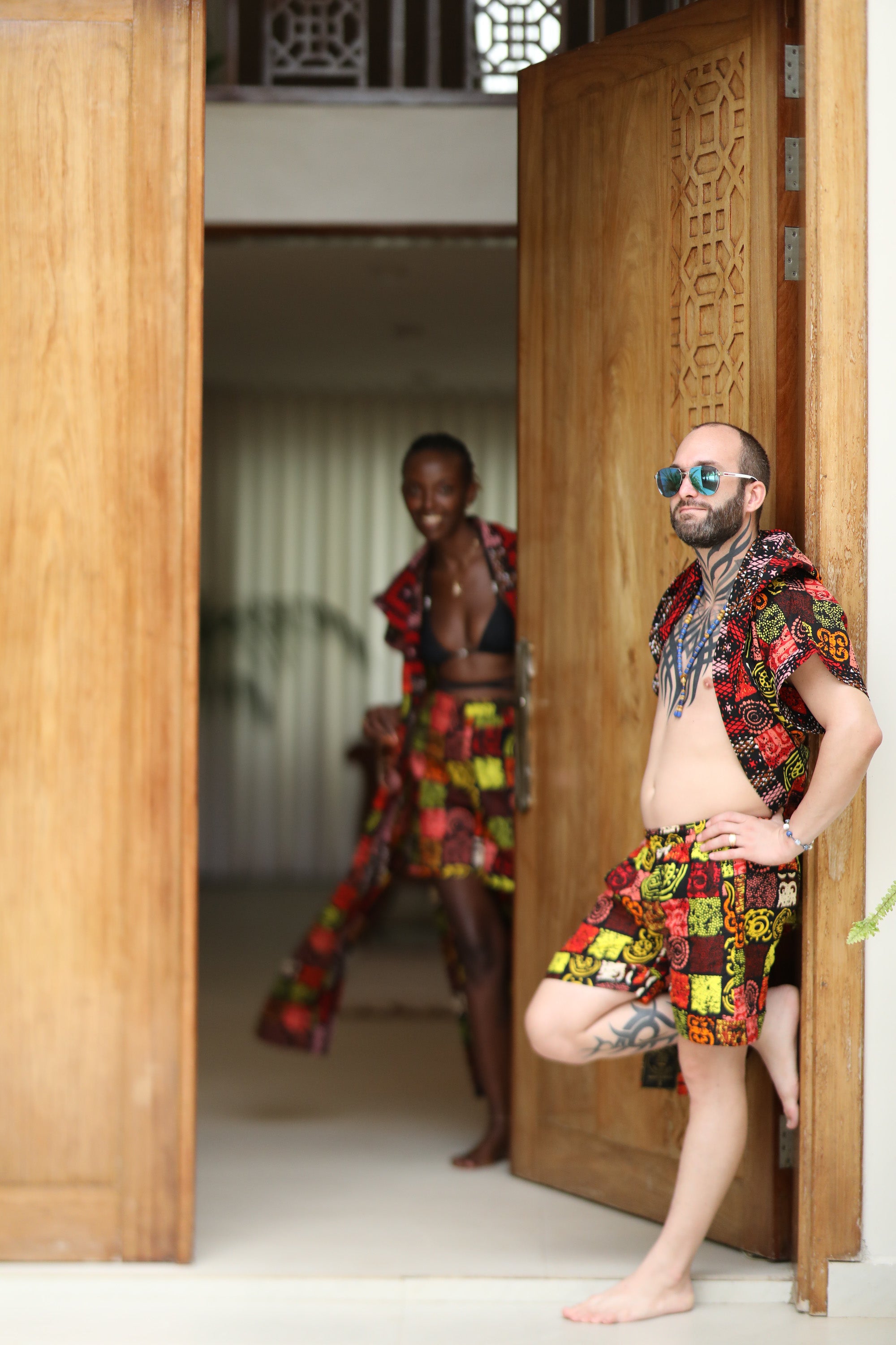 A couple styling Electric Safari African cover-up and shorts set in safari-inspired print – Zawadi Hat standing in a doorway with wooden panels.