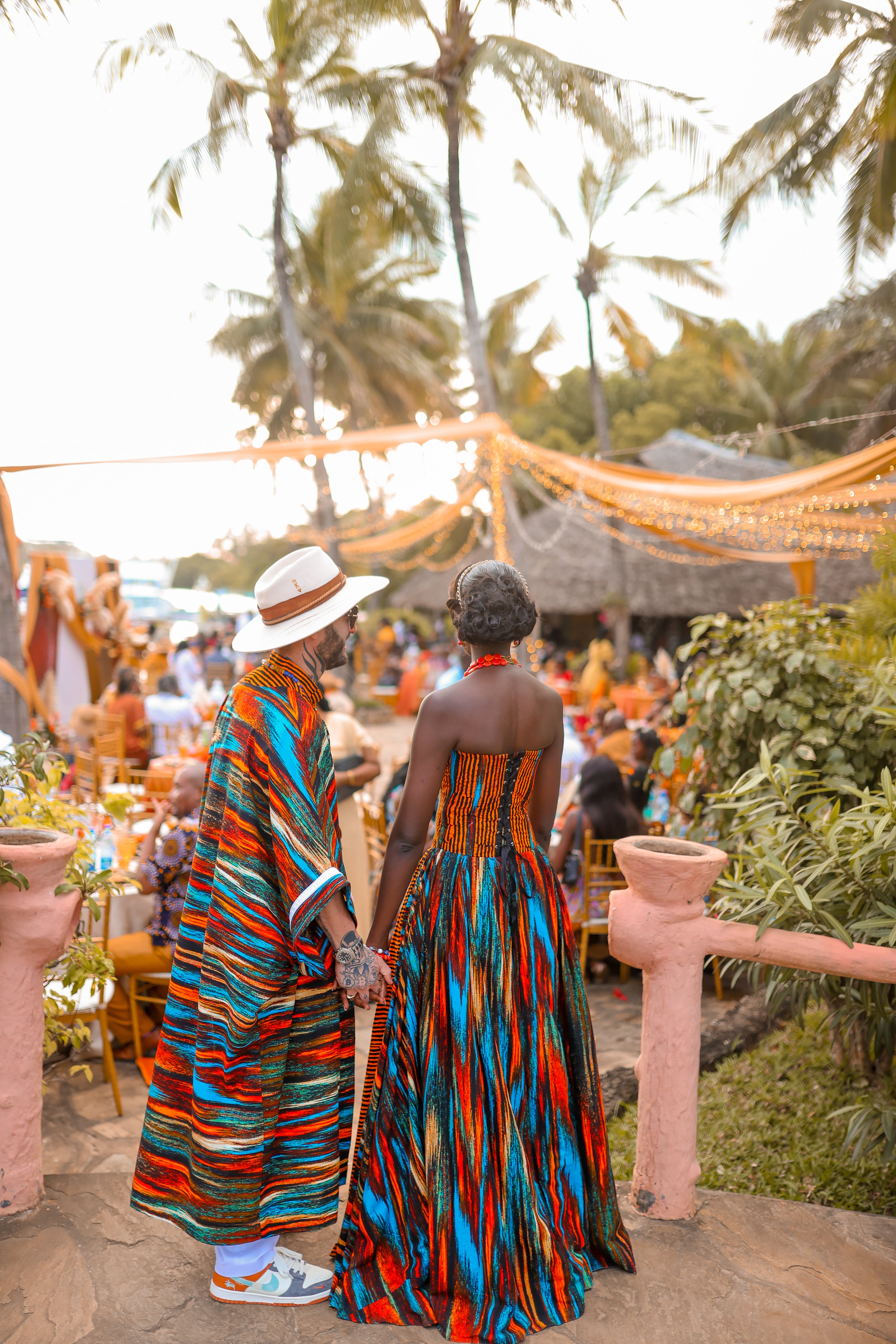 Zawadi Hat Fashion founders in colorful traditional attire walking together at their outdoor wedding with palm trees and guests in the background.